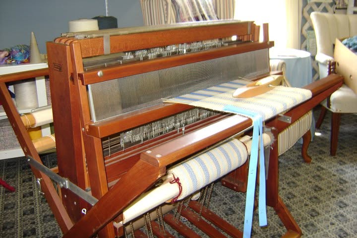 Wooden floor loom with blue-and-cream cloth and a wooden shuttle
