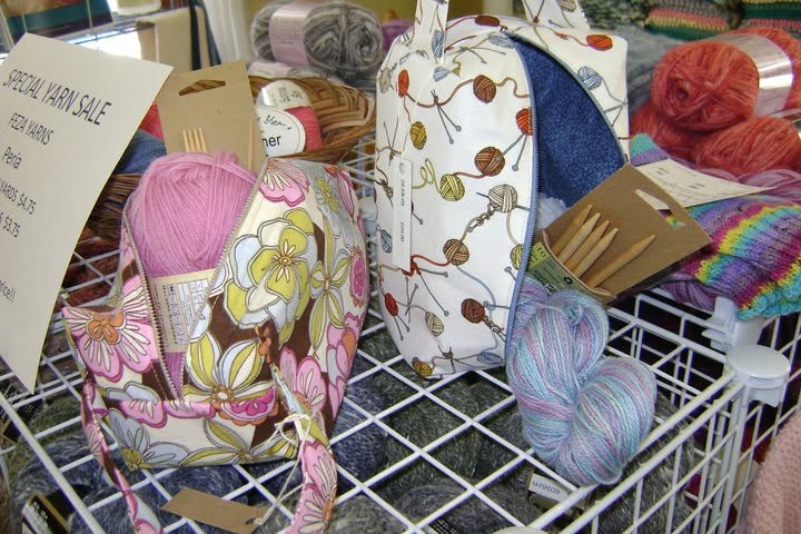 Floral and yarn-patterned project bags beside yarn skeins and wooden needles